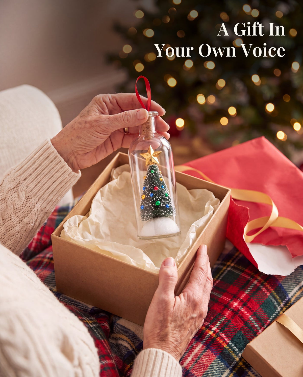 A woman's hands lifting the bottle out of a kraft gift box with tissue paper, on a plaid blanket, with red wrapping paper beside it.