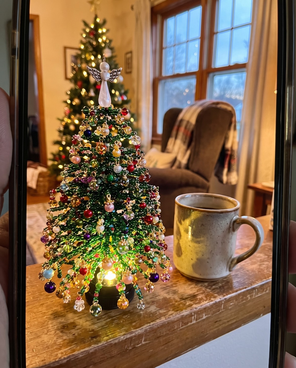 A finished beaded tree lit on a wooden table with the family Christmas tree behind it